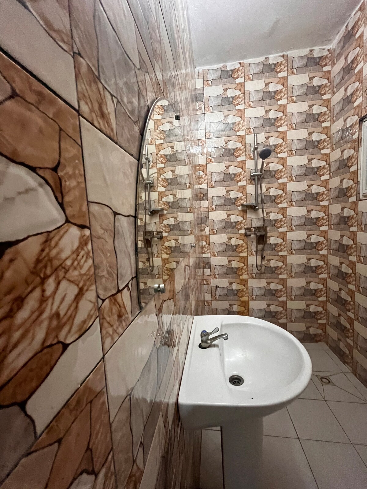 The bathroom features a wall-mounted shower with adjustable fixtures and a circular mirror. The walls are adorned with a unique patterned tile design in shades of brown and beige, complemented by a compact pedestal sink positioned adjacent to the shower.