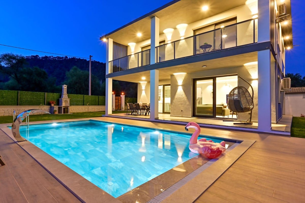 A modern villa is presented under evening lights, featuring a spacious pool with clear blue water. A pink inflatable float rests on the water's surface. The structure has multiple balconies, and a cozy seating area is visible through large glass doors, providing an inviting view of the garden.