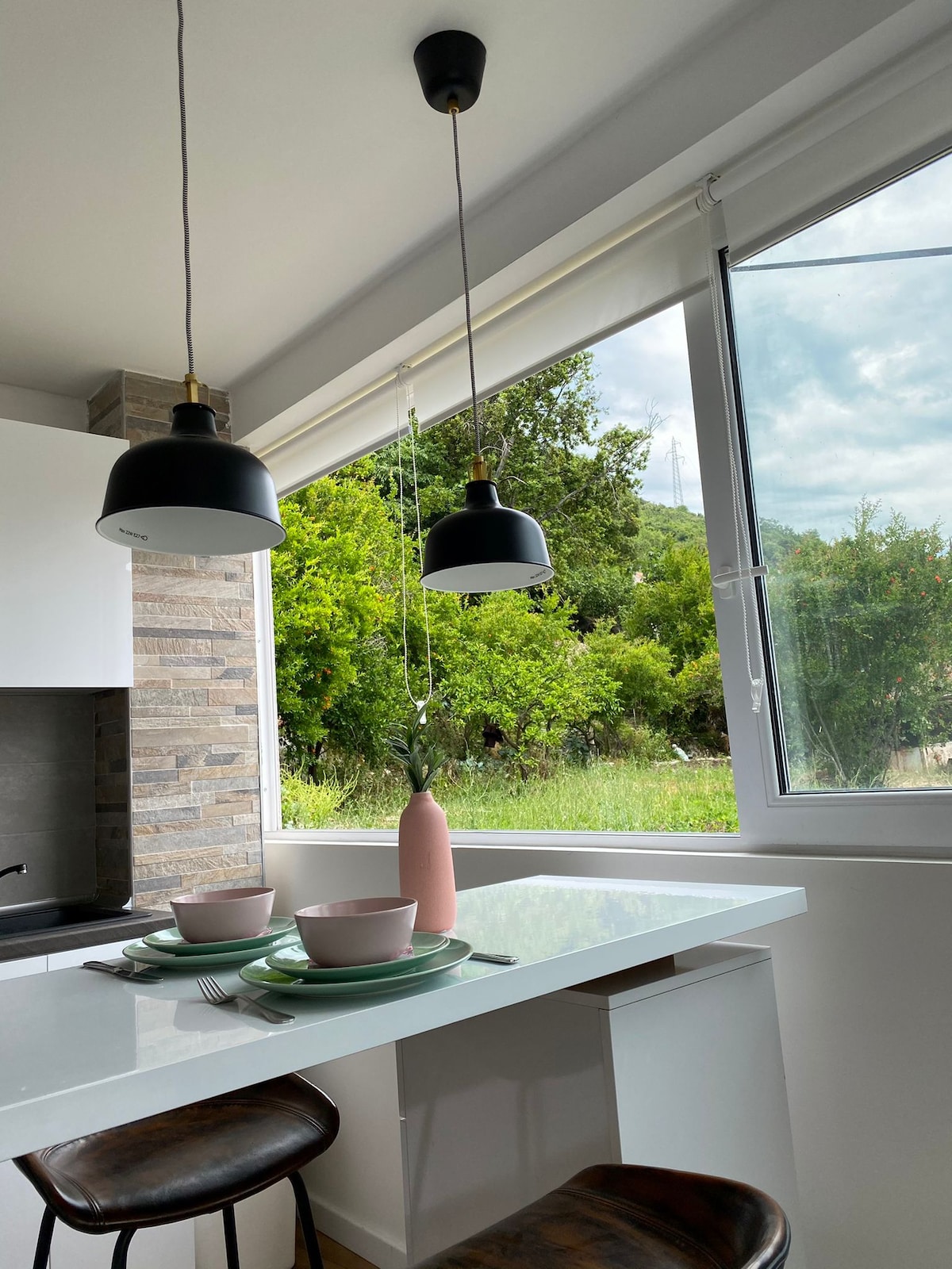 A modern kitchen space features a white countertop with two place settings, each including a plate and bowl. Two pendant lights hang above the counter, and a large window offers views of greenery outside, while natural light brightens the area.