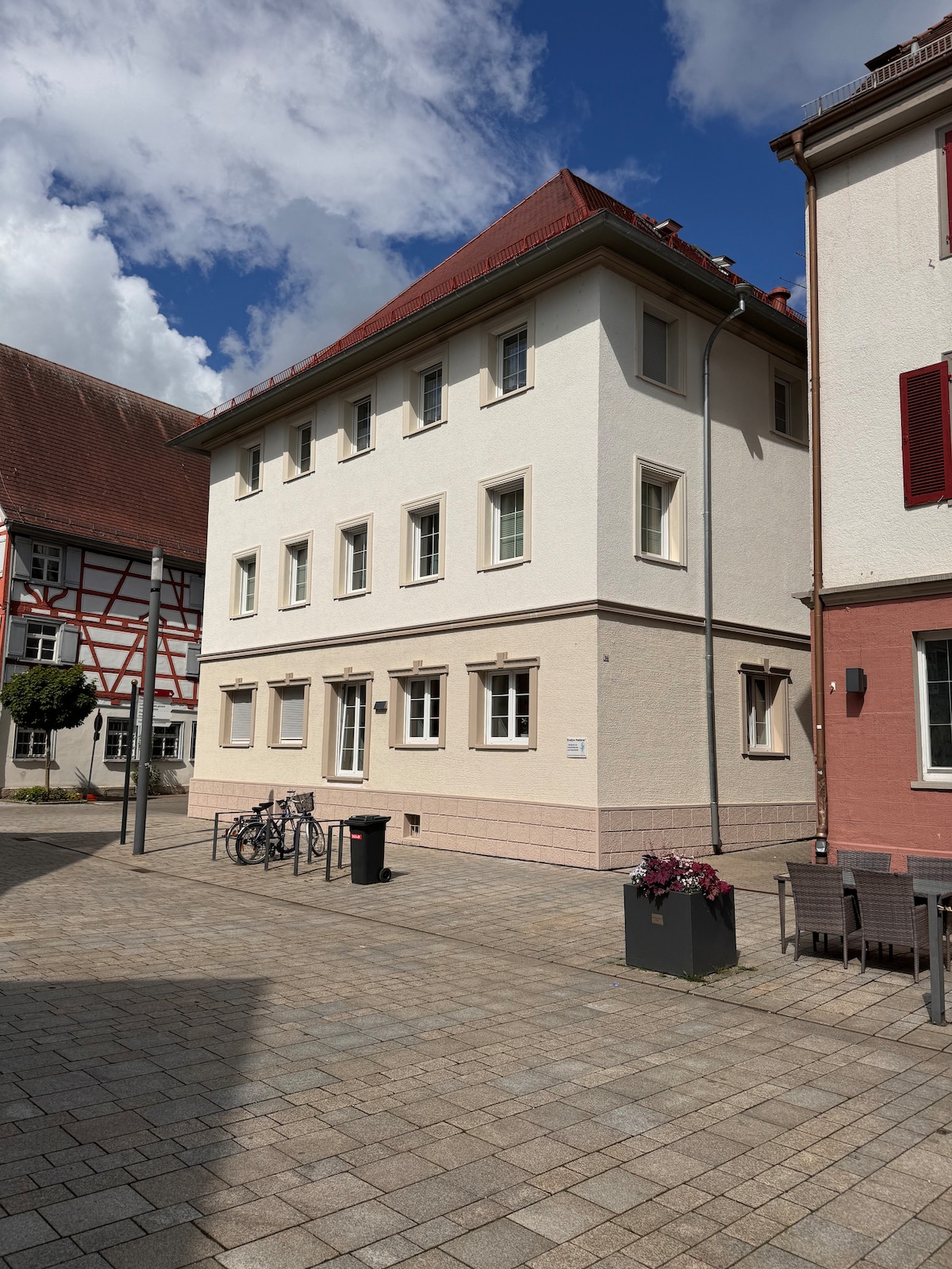 The image depicts a three-story building characterized by a clean white exterior and a red sloped roof. Windows are evenly spaced along the sides, providing natural light. Nearby, outdoor seating is arranged around planters filled with flowers, and bicycle racks are visible along the pathway.