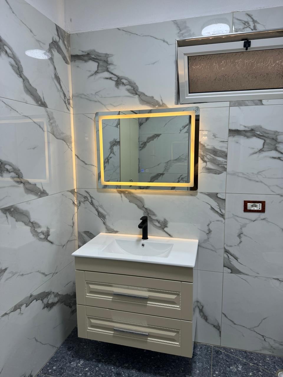 A modern bathroom features elegant marble-patterned walls and a sleek vanities. A well-lit mirror is framed with warm lighting, enhancing visibility. A window offers natural light, complementing the clean design and functional storage space beneath the sink.