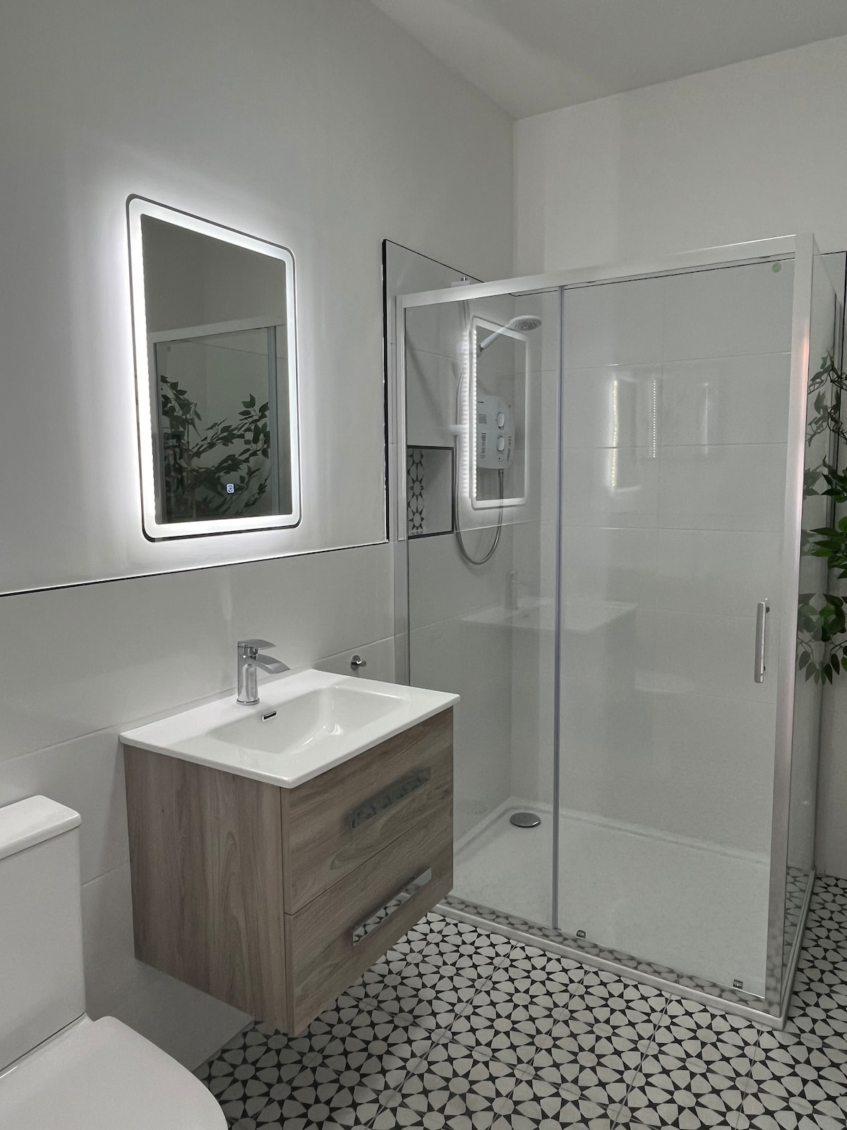 The spacious bathroom features a modern design with a large walk-in shower enclosed in glass. A stylish wooden vanity holds a sleek sink, and a backlit mirror adds illumination. Distinctive patterned tiles cover the floor, enhancing the contemporary feel.