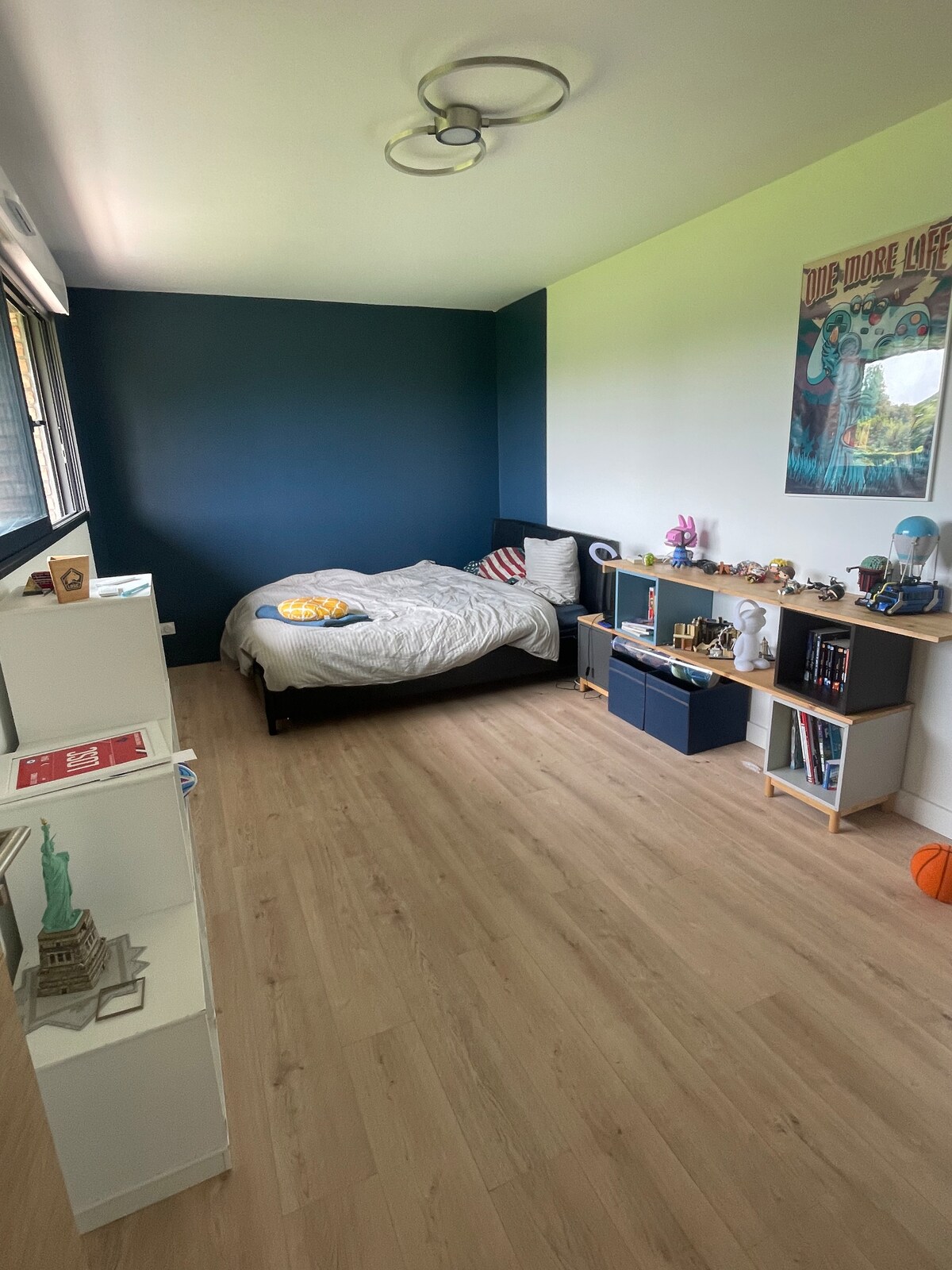 A well-lit bedroom features a spacious bed with a light-colored blanket and decorative pillows. A dark wall contrasts with the lighter walls, and shelving units display various items. A window allows natural light to fill the room, contributing to the serene atmosphere.
