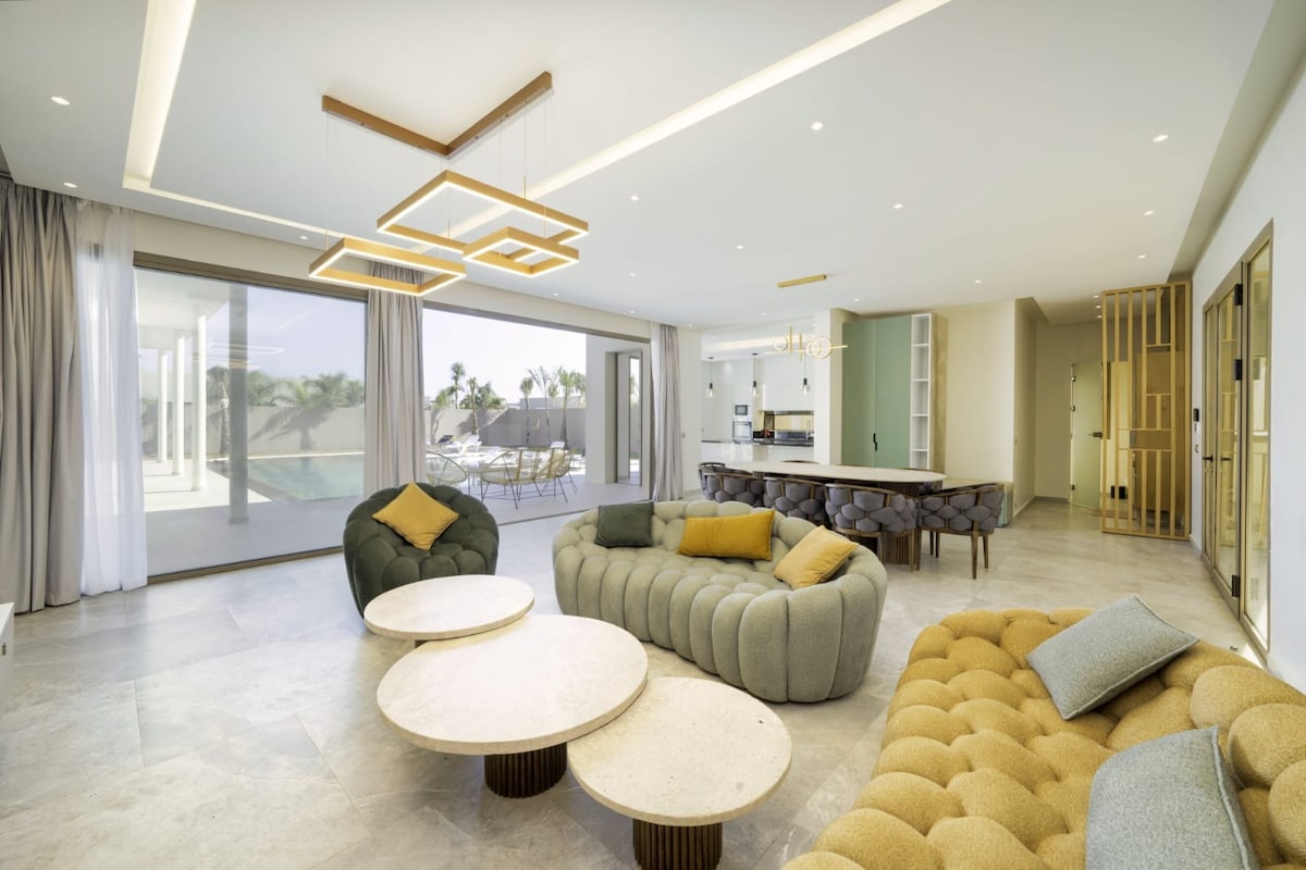 A spacious living area features modern furniture with two large, textured sofas in soft colors and two round coffee tables at the center. Natural light floods the room through expansive glass doors, leading to an outdoor space. Unique lighting fixtures add interest to the ceiling.