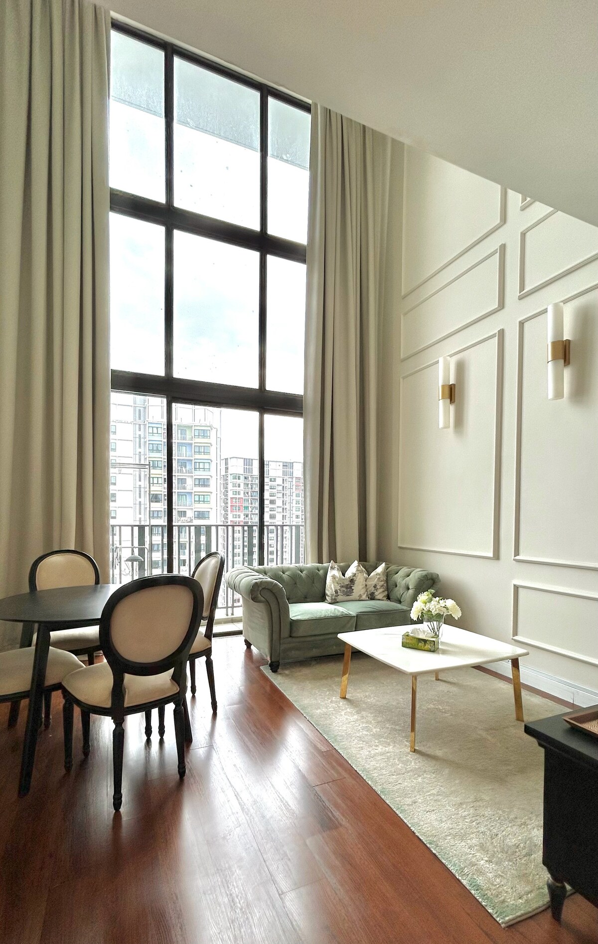 A stylish living area is highlighted by tall windows, allowing ample natural light to illuminate the space. A round dining table with four chairs complements a cozy green sofa, which is adorned with decorative cushions. A coffee table centers the room, placed on a soft area rug.