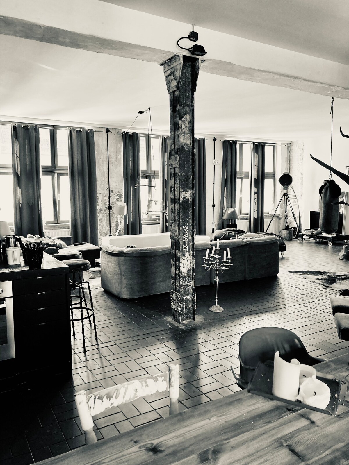 The open living area showcases high ceilings and exposed brickwork, with large windows allowing natural light to fill the space. A variety of seating options, including three couches, can be seen, along with decorative elements that reflect the former factory setting.