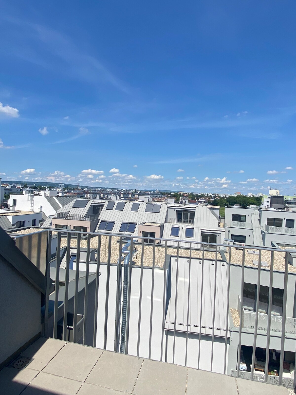 A rooftop view captures a vibrant skyline under a clear blue sky. Neatly arranged rooftops with solar panels can be seen, along with distant hills, creating a spacious atmosphere. The scene conveys a sense of openness and connection to the surrounding environment.
