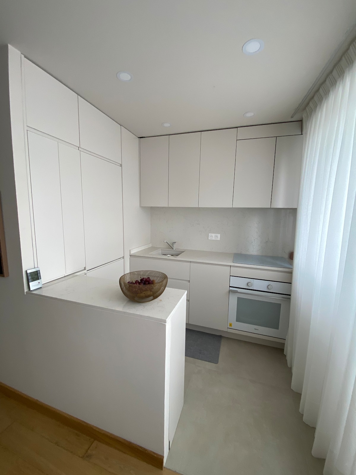 A sleek, modern kitchenette is presented, featuring white cabinetry and a light countertop. A small bowl of fruit is placed on the counter. Natural light filters in through sheer curtains, enhancing the airy feel of the space.