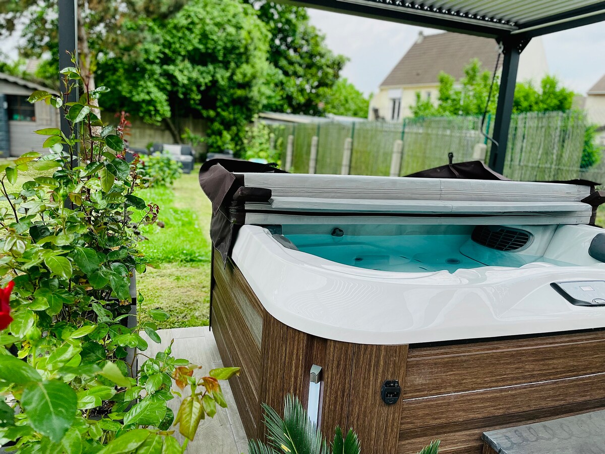A hot tub is situated under a covered area, featuring a sleek, modern design with light blue water visible inside. Green foliage surrounds the hot tub, providing a natural backdrop, while grass and trees are seen in the distance.