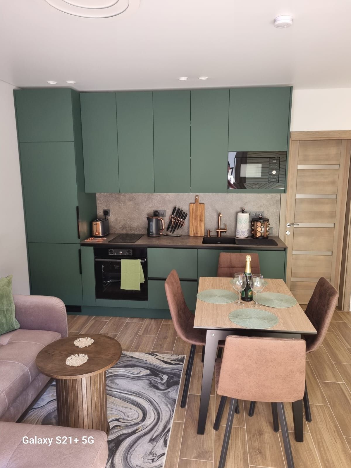 A modern kitchen and dining area feature sage green cabinetry and a sleek countertop. An oven, stovetop, and microwave are included. A wooden dining table is paired with four upholstered chairs. A cozy sofa is positioned nearby, creating a functional and inviting space.