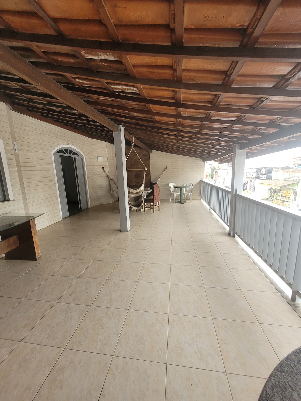An expansive covered balcony features a tiled floor and wooden ceiling. A small table and several chairs are positioned along one side, while a decorative hammock is displayed nearby. The space is open and well-lit, providing views to the outdoors.