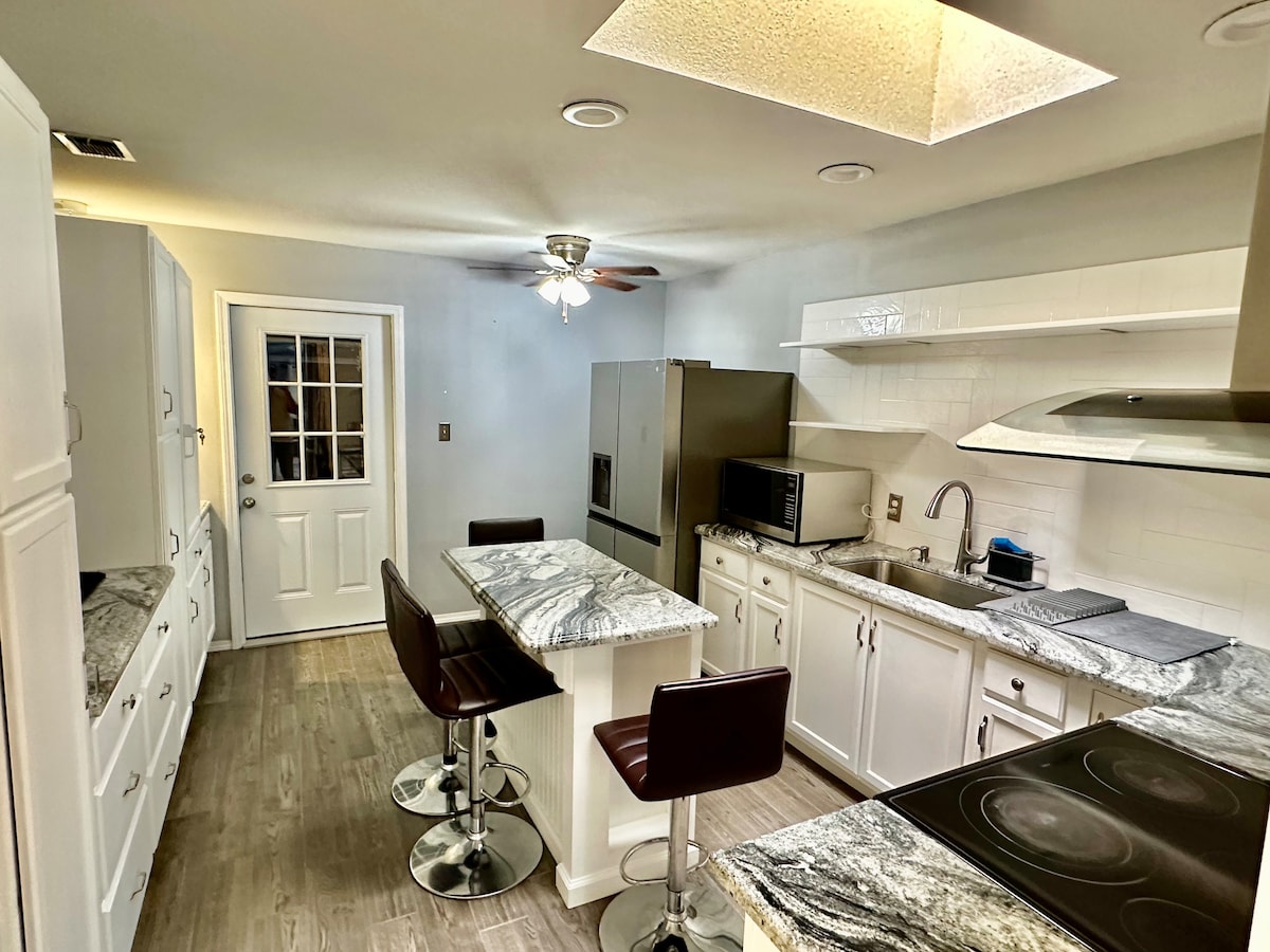 A modern kitchen features white cabinetry with granite countertops, complemented by stainless steel appliances. An island with seating for two is highlighted, alongside a door leading to an outdoor area. Natural light is provided by overhead lights and an oversized skylight.