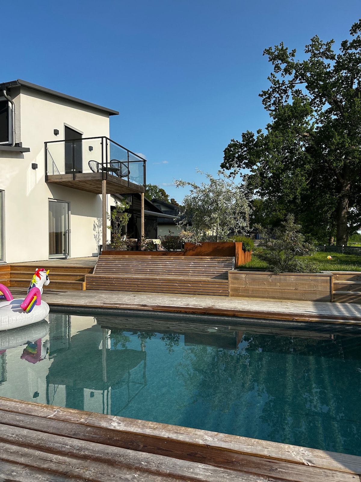 A new pool constructed in 2024 is surrounded by wooden decking, providing a serene outdoor space. The house features a modern design with large glass doors, and a balcony overlooks the tranquil setting punctuated by greenery and landscaped areas.
