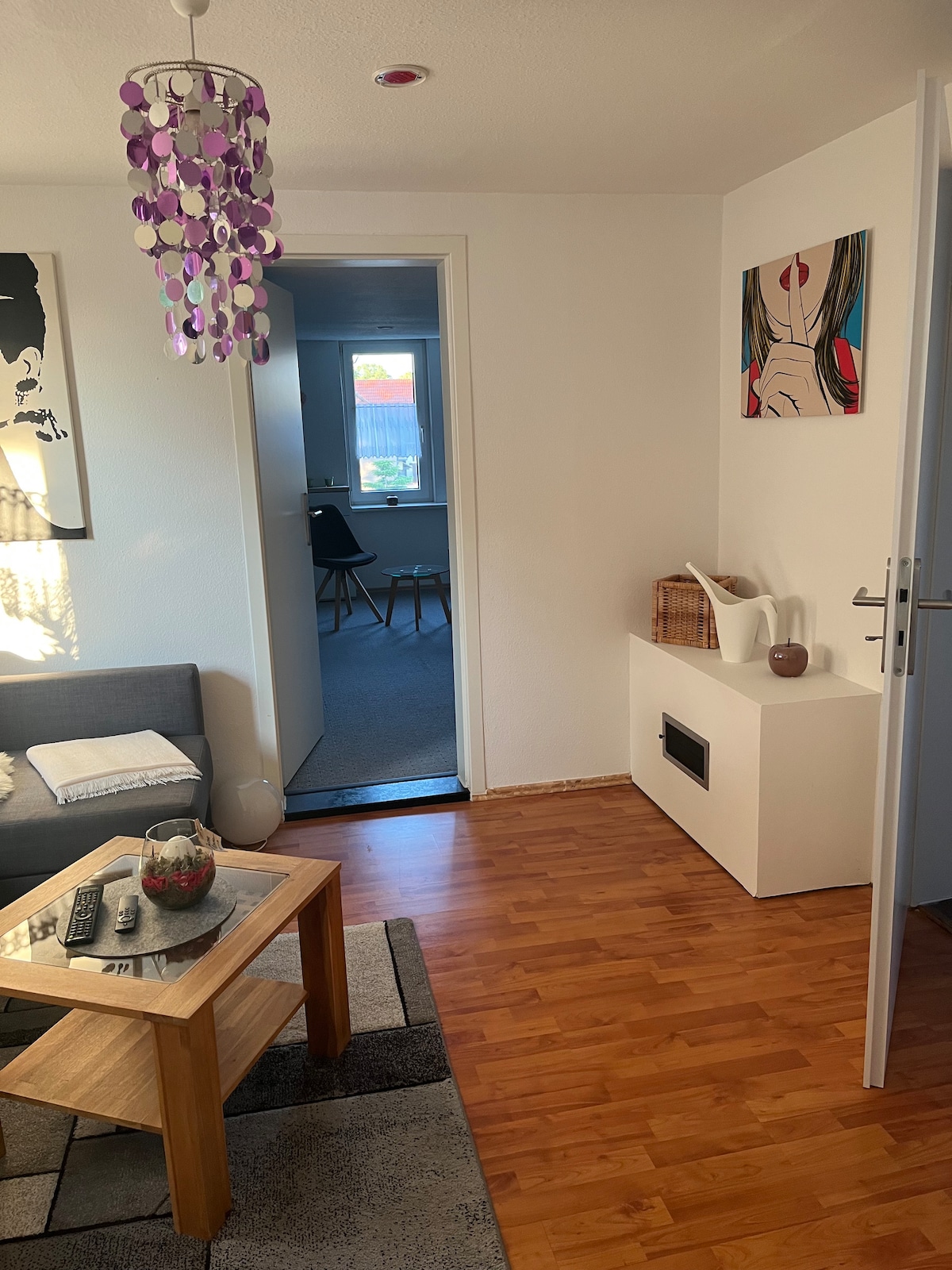 A living area features a cozy gray sofa beside a wooden coffee table with a decorative bowl. A small, multi-colored chandelier hangs overhead. A doorway leads to a well-lit room with minimal furnishings, while a contemporary art piece adorns the wall.