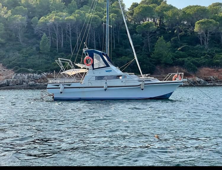 A sailboat is anchored in calm waters, surrounded by lush green trees. The boat features a canopy over the cockpit and life rings are visible on either side. The tranquil setting emphasizes a connection with nature.