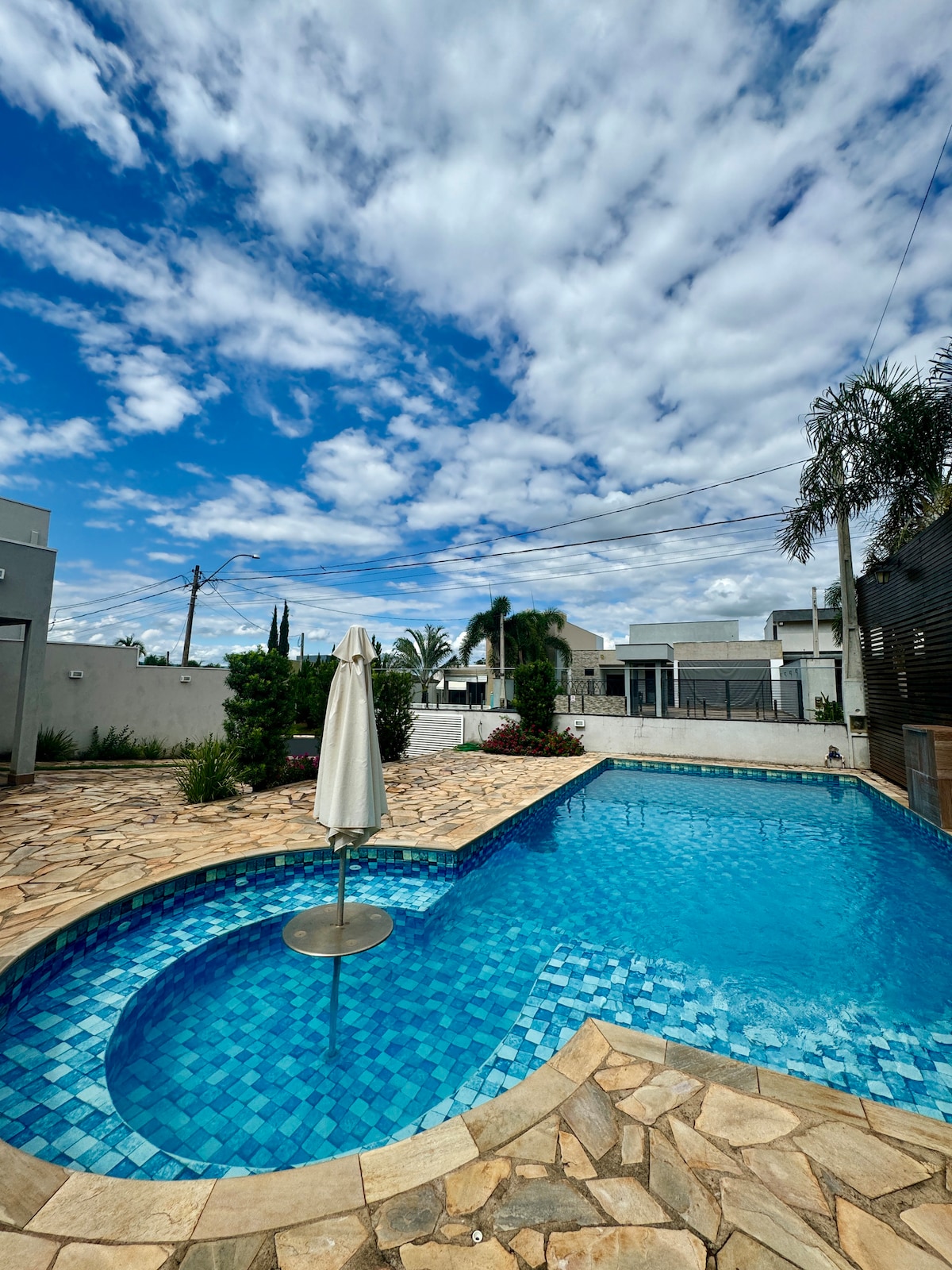 A spacious outdoor swimming pool with vibrant blue tiles is featured, surrounded by a stone patio. A white umbrella provides shade over the pool area. Lush greenery and residential structures are visible in the background under a partly cloudy sky.