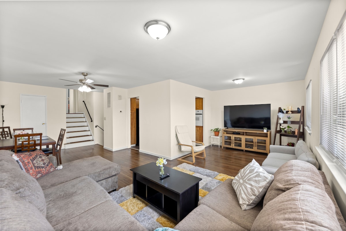 A spacious living area features a large sectional sofa set on a soft rug, complemented by a coffee table. Natural light streams through multiple windows, illuminating the room, which includes a flat-screen TV, a cozy armchair, and a dining table nearby.