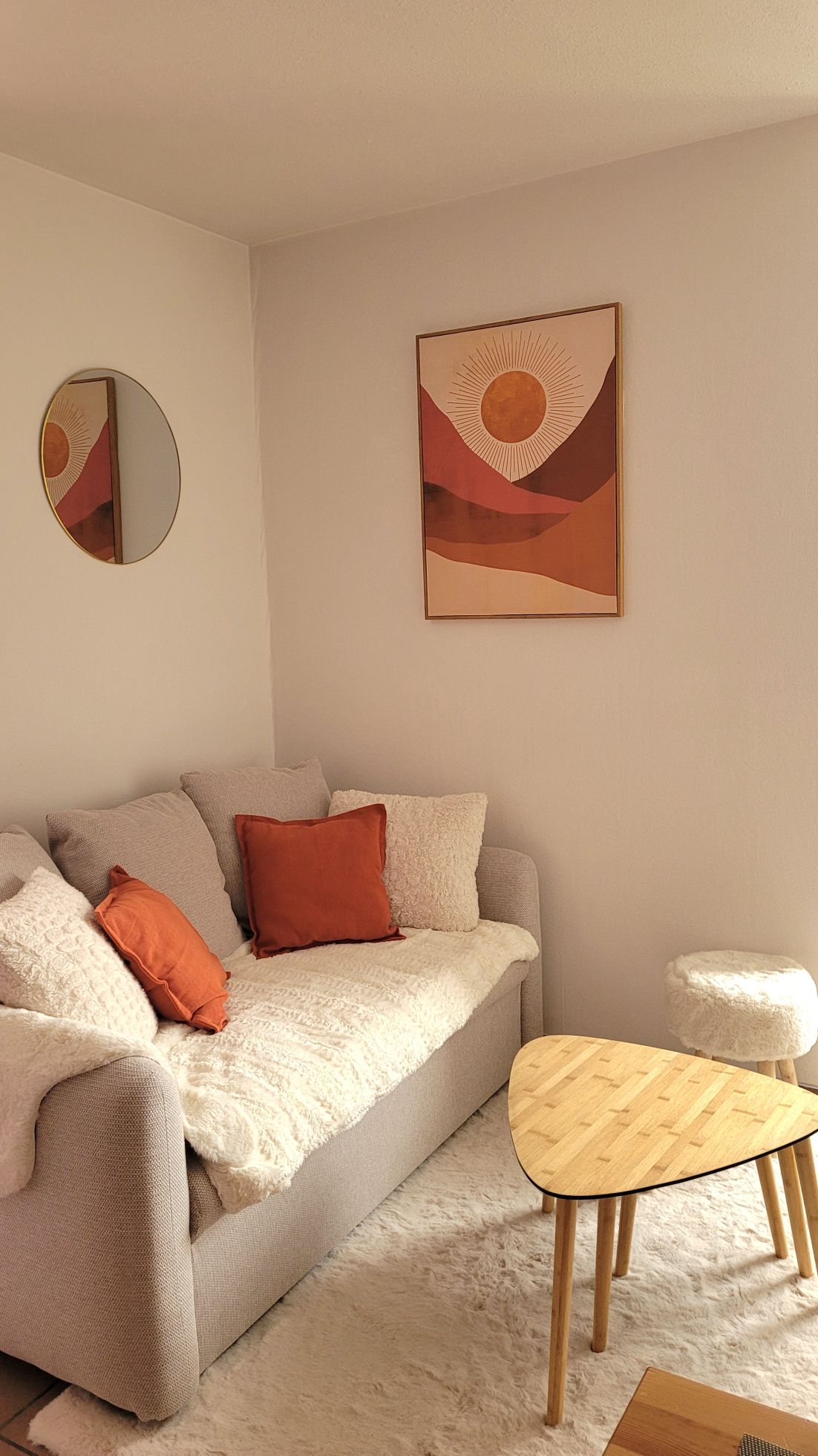 A comfortable seating area features a light gray sofa adorned with decorative cushions in earthy tones. A round mirror is mounted on the wall, reflecting natural light. A small, unique wooden table and a textured, round stool complement the cozy ambiance.