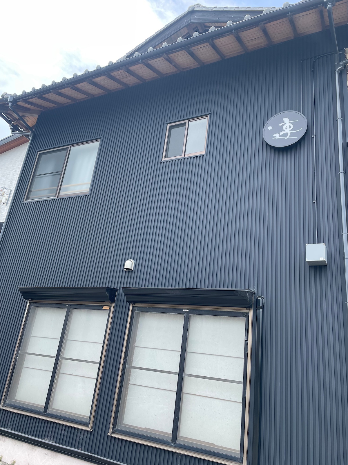 A two-story building is shown with a dark corrugated exterior. The upper floor features a single window, while the lower floor has two large windows separated by a visible sign. The roof showcases traditional Japanese architecture with wooden eaves.