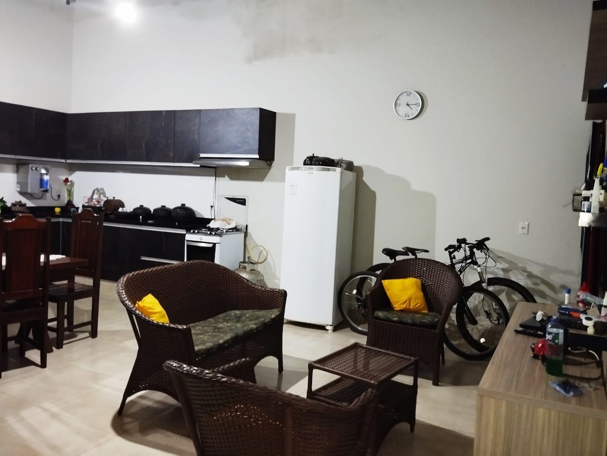 The open living area features a comfortable seating arrangement with wicker chairs and a small coffee table. A dining table is situated nearby. A refrigerator stands against a wall, while two bicycles are visible, highlighting the space's practicality.