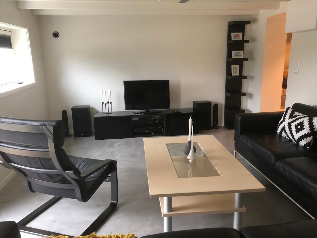 A modern living area is presented with a large flat-screen TV positioned atop a sleek entertainment unit. Comfortable seating includes a black leather couch and a contemporary black chair. A light coffee table features a central glass panel, with decorative candles placed on top.