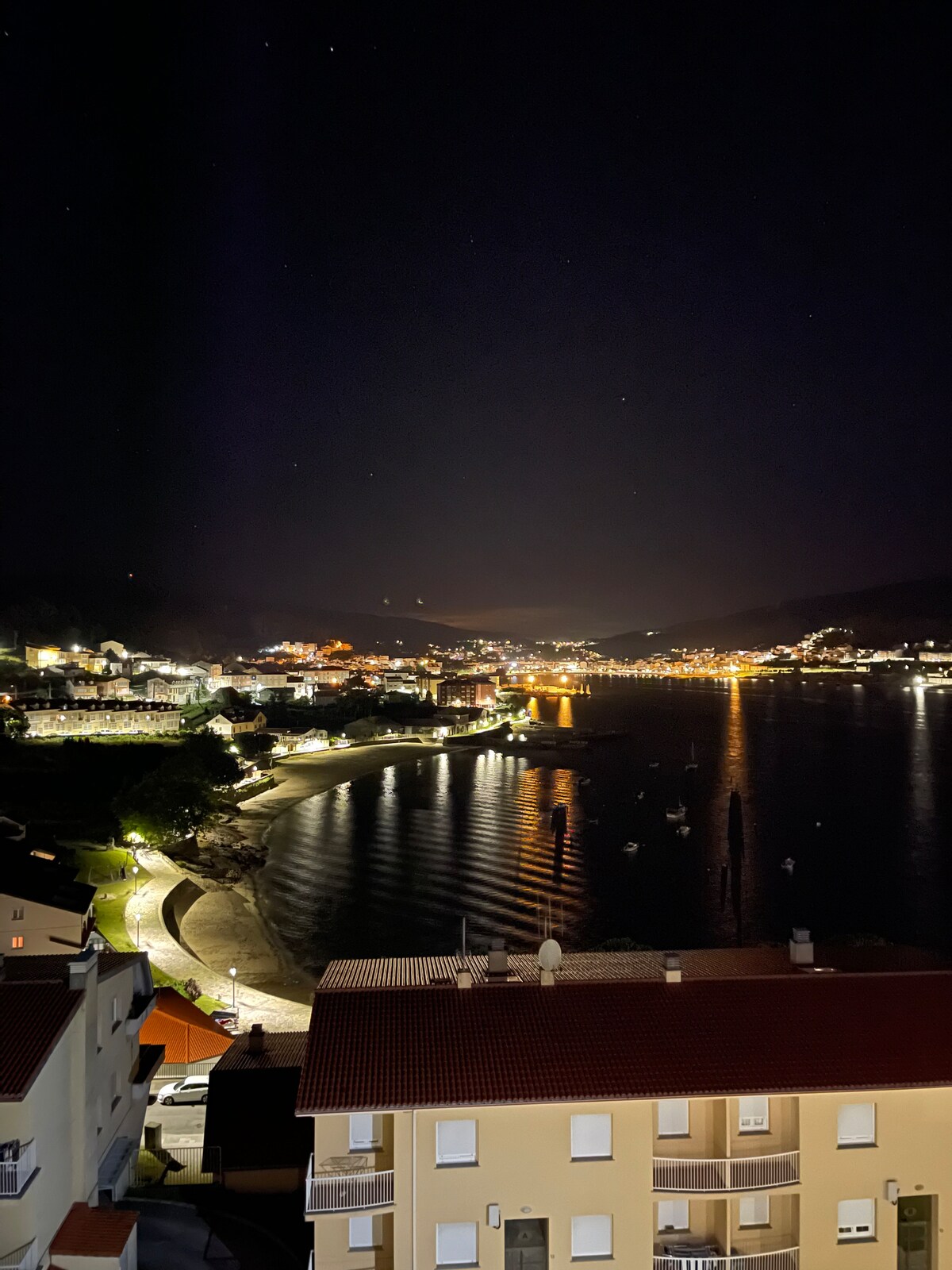 A nighttime view reveals a serene coastal town illuminated by soft lights. The shimmering water reflects the golden hues, while boats are anchored peacefully. A gentle curve of the shoreline is visible, bordered by lit pathways and charming buildings.