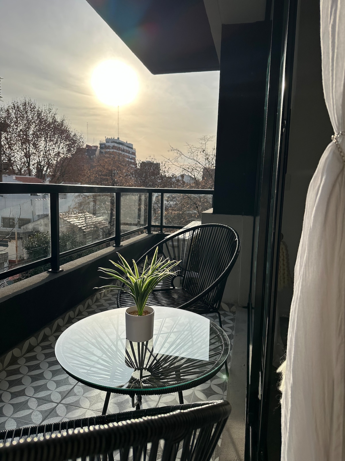 A balcony is presented with a round glass table and two black woven chairs, complemented by a small potted plant. The expansive view features a sunset illuminating the sky, casting a warm glow over the surrounding buildings and trees.