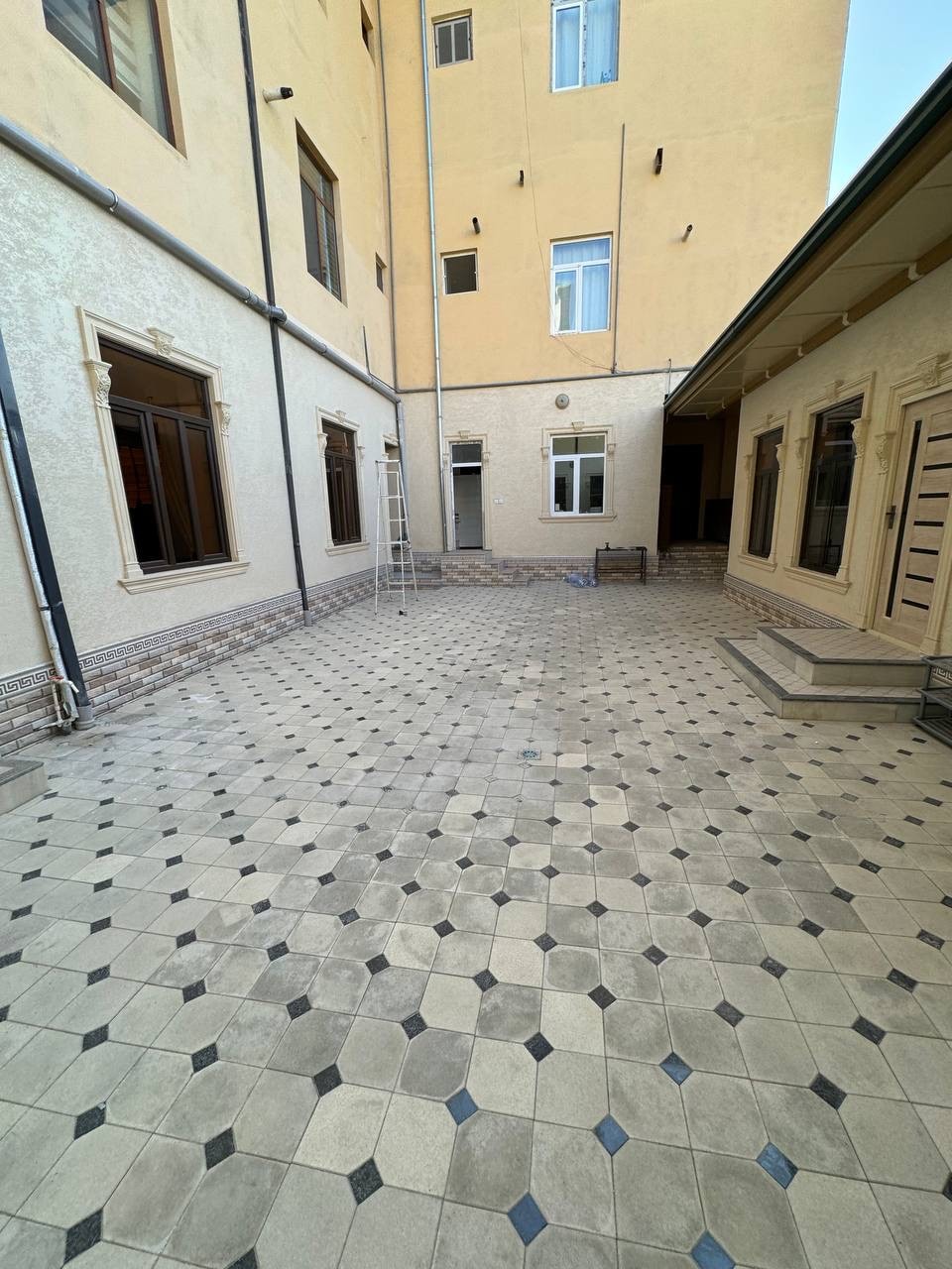 A spacious courtyard is showcased, featuring tiled flooring with a geometric pattern in neutral tones. Surrounding walls are light-colored, with multiple windows allowing natural light to enter. A ladder is positioned against one wall, indicating ongoing maintenance or accessibility.
