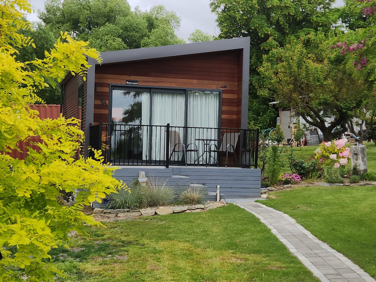 A modern guest house is surrounded by a lush garden, featuring a private deck with two chairs. Large glass doors provide access to the interior, which is framed by a sleek overhang. A stone pathway leads through the well-maintained lawn, enhancing the serene outdoor setting.