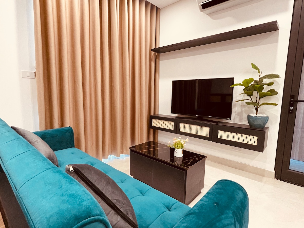A modern living area is highlighted by a plush teal sofa and a sleek black coffee table. A flat-screen TV is mounted on the wall, accompanied by a decorative potted plant. Floor-to-ceiling curtains create an elegant backdrop, adding to the room's contemporary style.