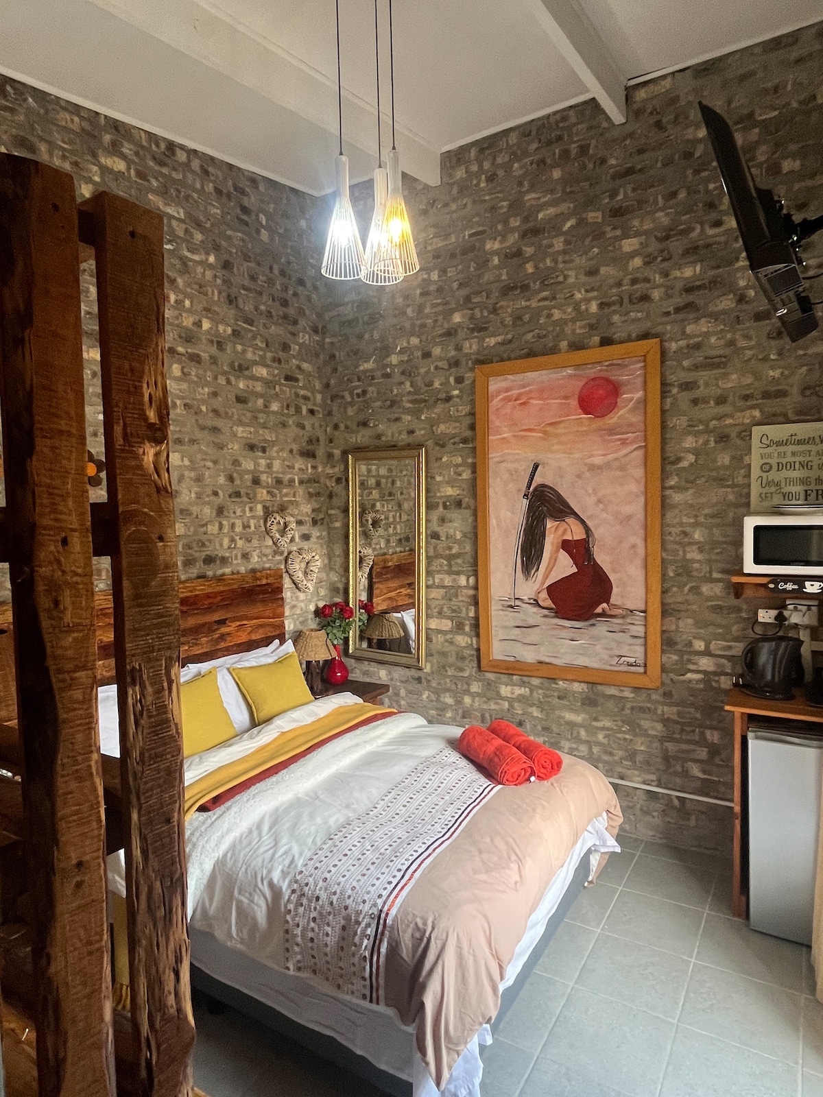 A warm and inviting bedroom features a queen bed adorned with neutral and vibrant linens. A large piece of wall art hangs above the bed, complemented by decorative mirrors. A coffee station is located nearby, alongside a small television mounted on the wall.