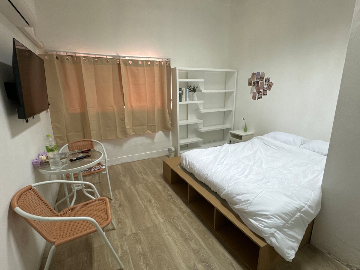 A bright and airy room features a spacious bed with white linens against a light wall. A small round table is accompanied by two chairs, and a shelf displays decorative items. Soft light enters through a window covered with beige curtains.