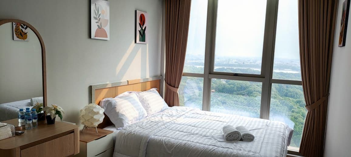 A bright bedroom features a spacious bed with neatly arranged linens, framed by large windows offering expansive views of greenery. A simple wooden dresser complements the decor, and fresh towels are neatly placed beside a small lamp on the bedside table.