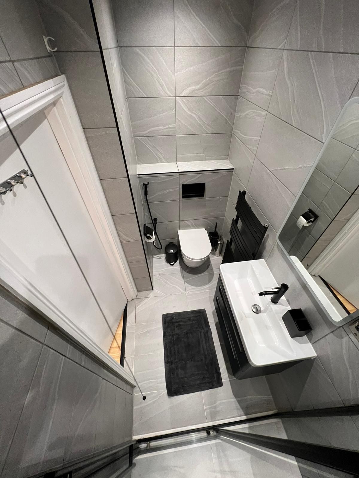 A modern bathroom features sleek gray tiles throughout, complemented by minimalistic fixtures. A wall-mounted mirror reflects the space, while a black mat is positioned in front of a stylish vanity with a basin. The shower area is enclosed with glass panels, maintaining an open feel.