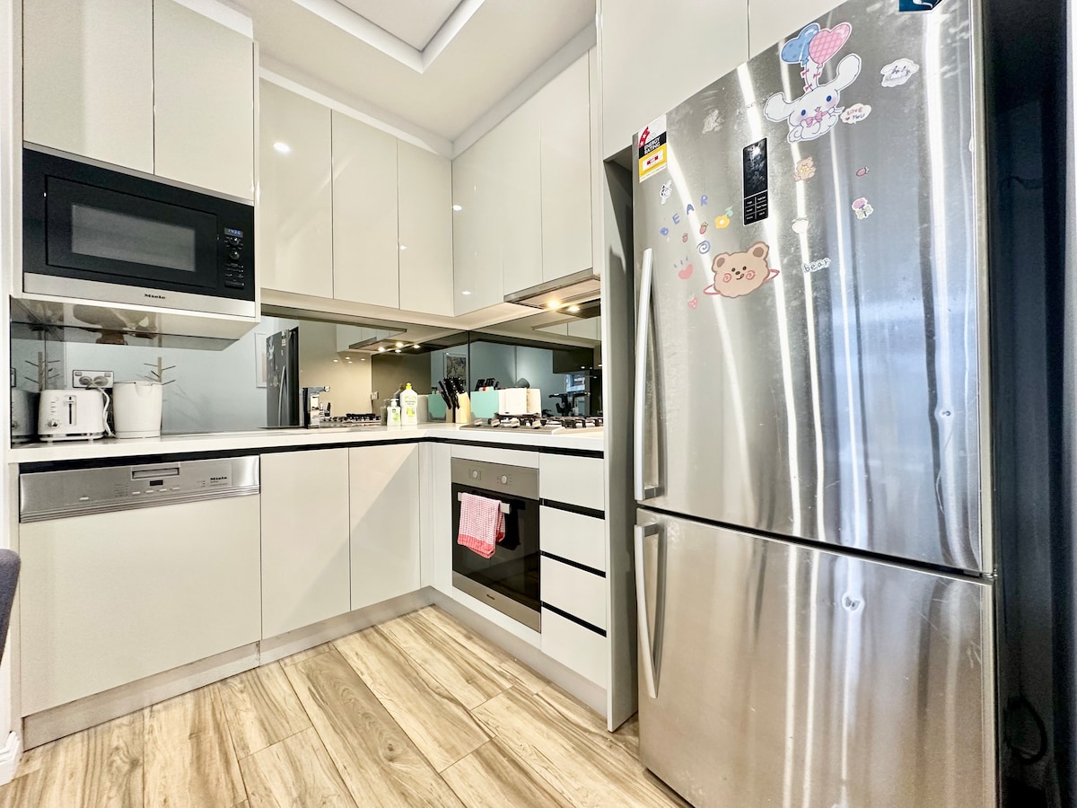 A modern kitchen is showcased with sleek cabinetry and stainless steel appliances. Essential equipment includes a microwave, oven, and gas stovetop. A refrigerator is adorned with playful magnets. Natural light enhances the space through a nearby window, complementing the wood-patterned flooring.