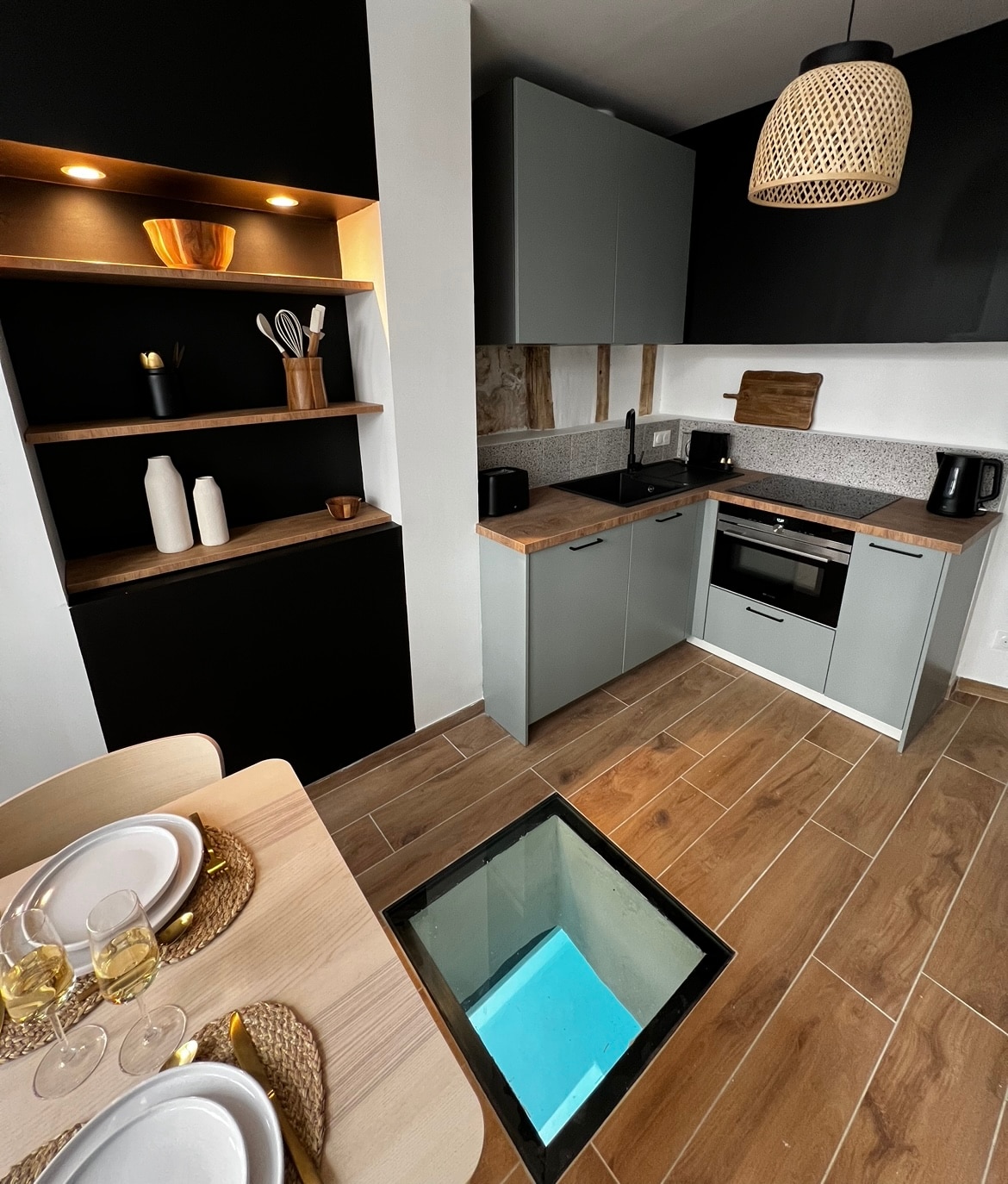 The well-equipped kitchen features modern cabinetry and a stylish light fixture. A glass panel reveals a glimpse of the pool below. The dining area includes a wooden table set for two, complemented by elegant tableware, creating a cozy space for meals.