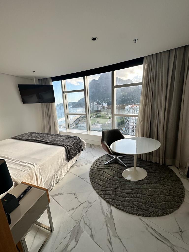 A guest room features a spacious bed with neutral bedding, complemented by a small round table and a modern chair. Large windows provide a view of the coastal landscape, while light-colored marble flooring adds a contemporary touch to the space.