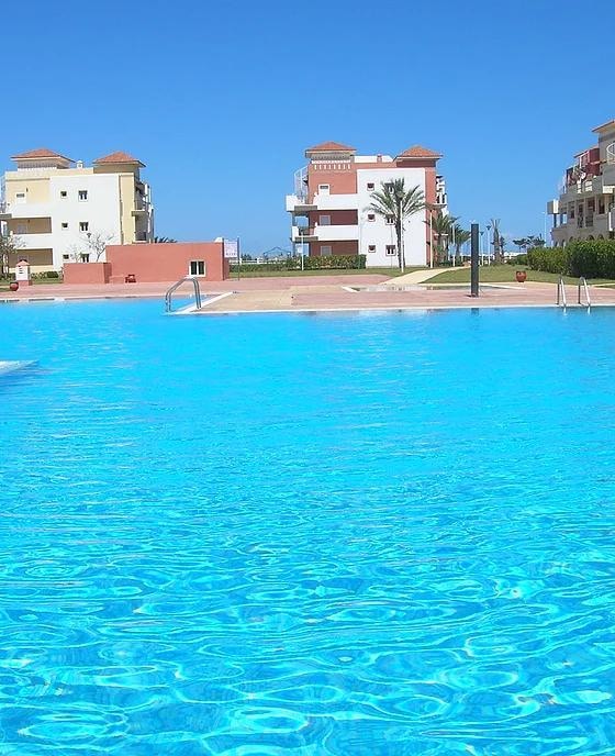 A spacious pool area is highlighted by clear turquoise water under a bright blue sky, surrounded by well-maintained gardens. Modern buildings with terracotta roofs are visible in the background, contributing to a serene and inviting setting.