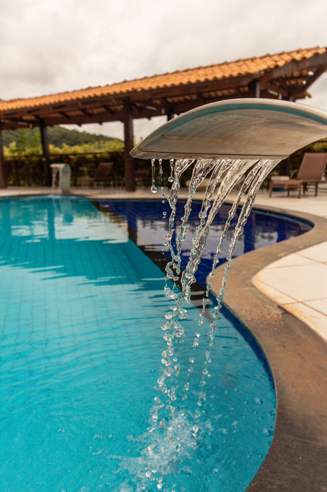 A serene swimming pool features a sleek water feature, cascading water into the sparkling blue water. A shaded seating area with lounge chairs offers relaxation beside the pool, surrounded by lush greenery and a rustic wooden structure with a tiled roof.
