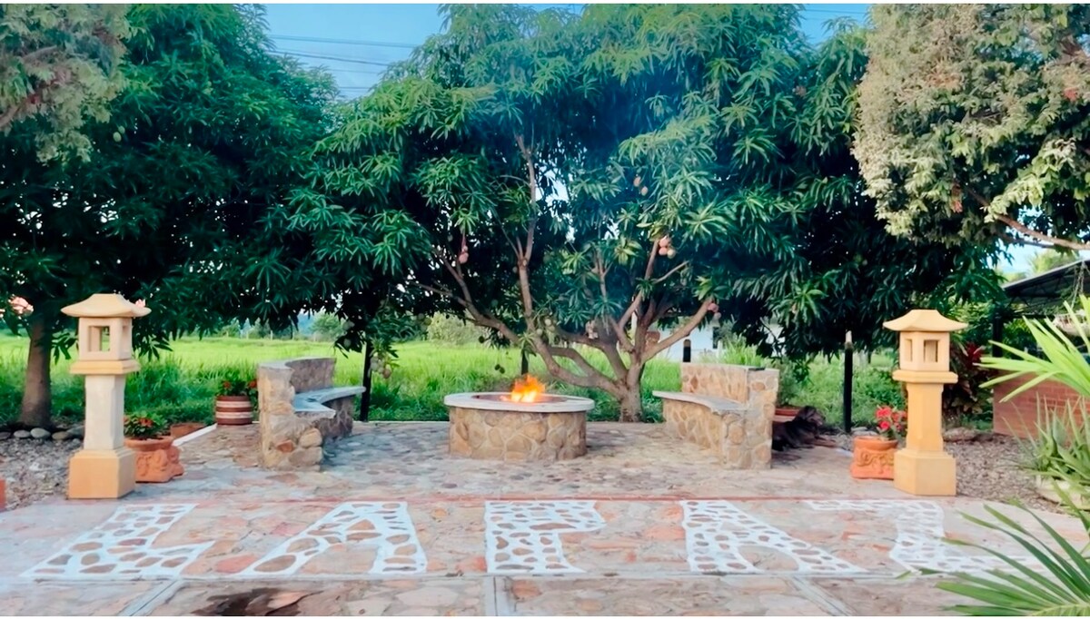 An inviting outdoor seating area is framed by lush trees and colorful greenery. Stone benches surround a circular fire pit, creating a central gathering space. Decorative lanterns are positioned on either side, complementing the natural stone path that leads to the seating area.