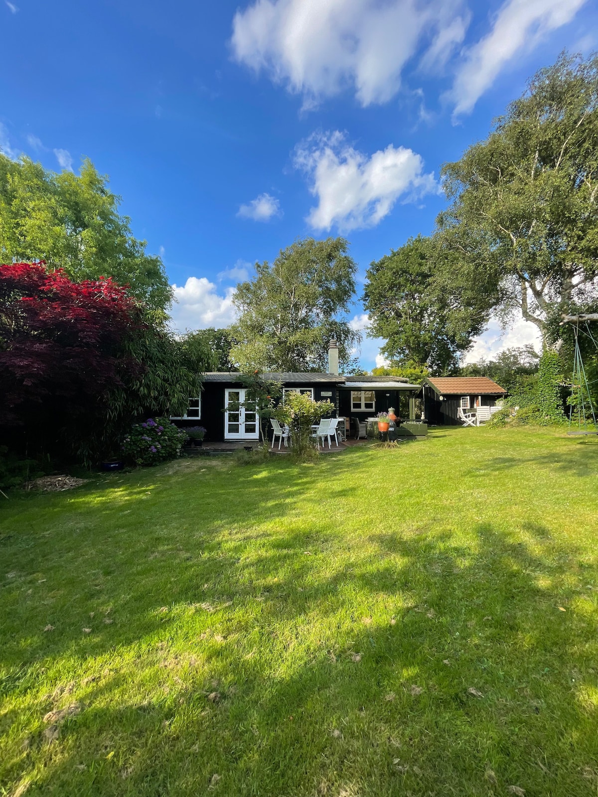The exterior of the summer house is depicted, surrounded by green grass and trees. A spacious garden area is shown, with a seating arrangement on a patio featuring several chairs and a table. The sky is bright with scattered clouds adding to the serene atmosphere.