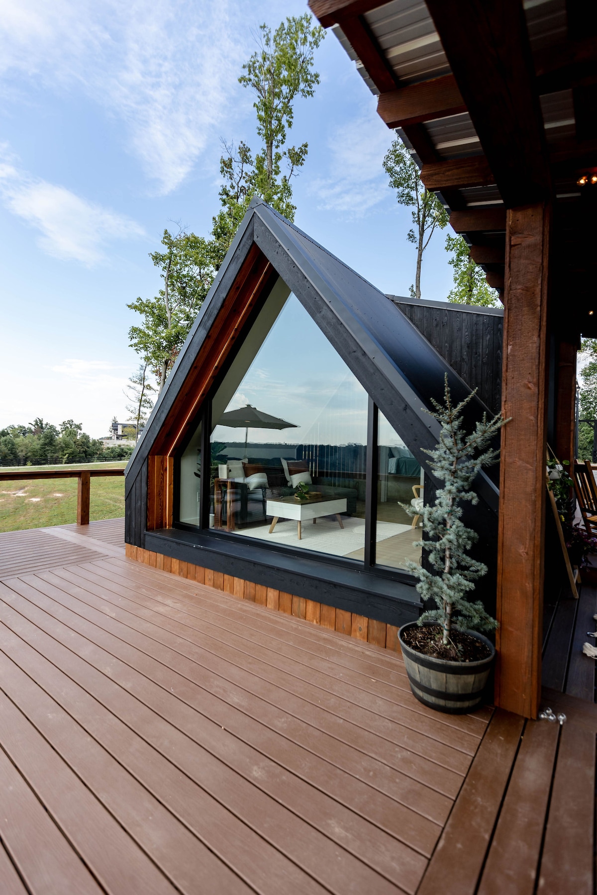 The exterior of an A-frame tiny home is shown, featuring a black roof and large glass windows. A wrap-around wooden deck is visible, with a potted evergreen plant placed beside the entrance. Natural surroundings and open space can be seen in the background.