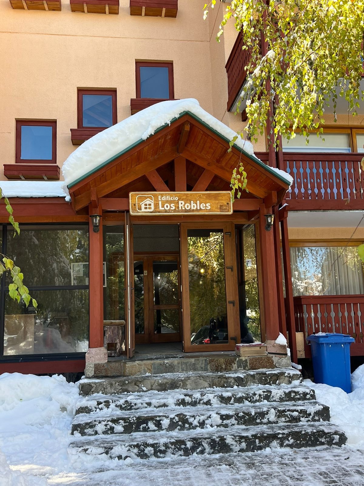 The entrance to Edificio Los Robles is depicted, featuring a wooden structure with a peaked roof. Snow blankets the steps leading up to large glass doors, while surrounding foliage adds a touch of greenery to the winter scene.