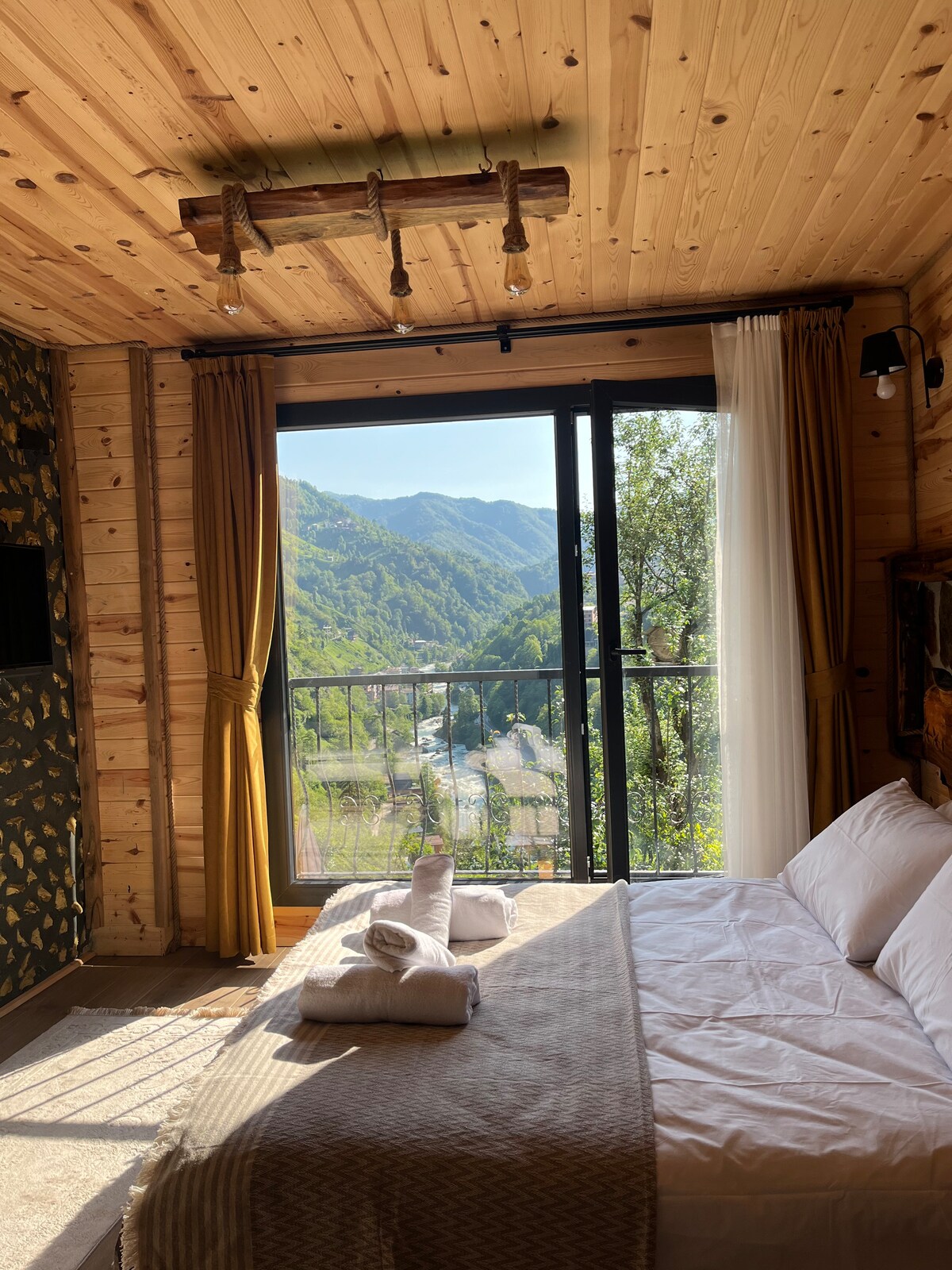 A sunlit bedroom features a spacious bed with neatly arranged towels. Floor-to-ceiling windows provide a view of the mountains, framed by light, flowing curtains. Warm wooden walls enhance the serene atmosphere, and soft lighting fixtures are mounted on a rustic beam above.