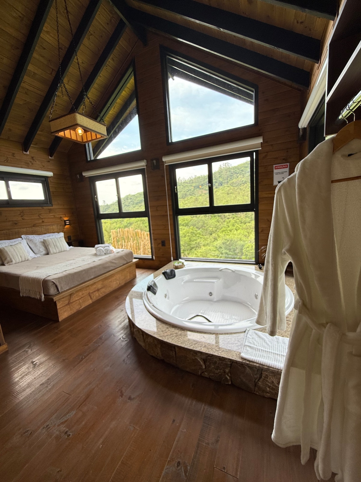 A wooden interior features a spacious bed beside a large window with scenic views. A round hot tub, framed by stone, is positioned prominently. A plush robe hangs nearby, emphasizing the relaxing atmosphere of the room.