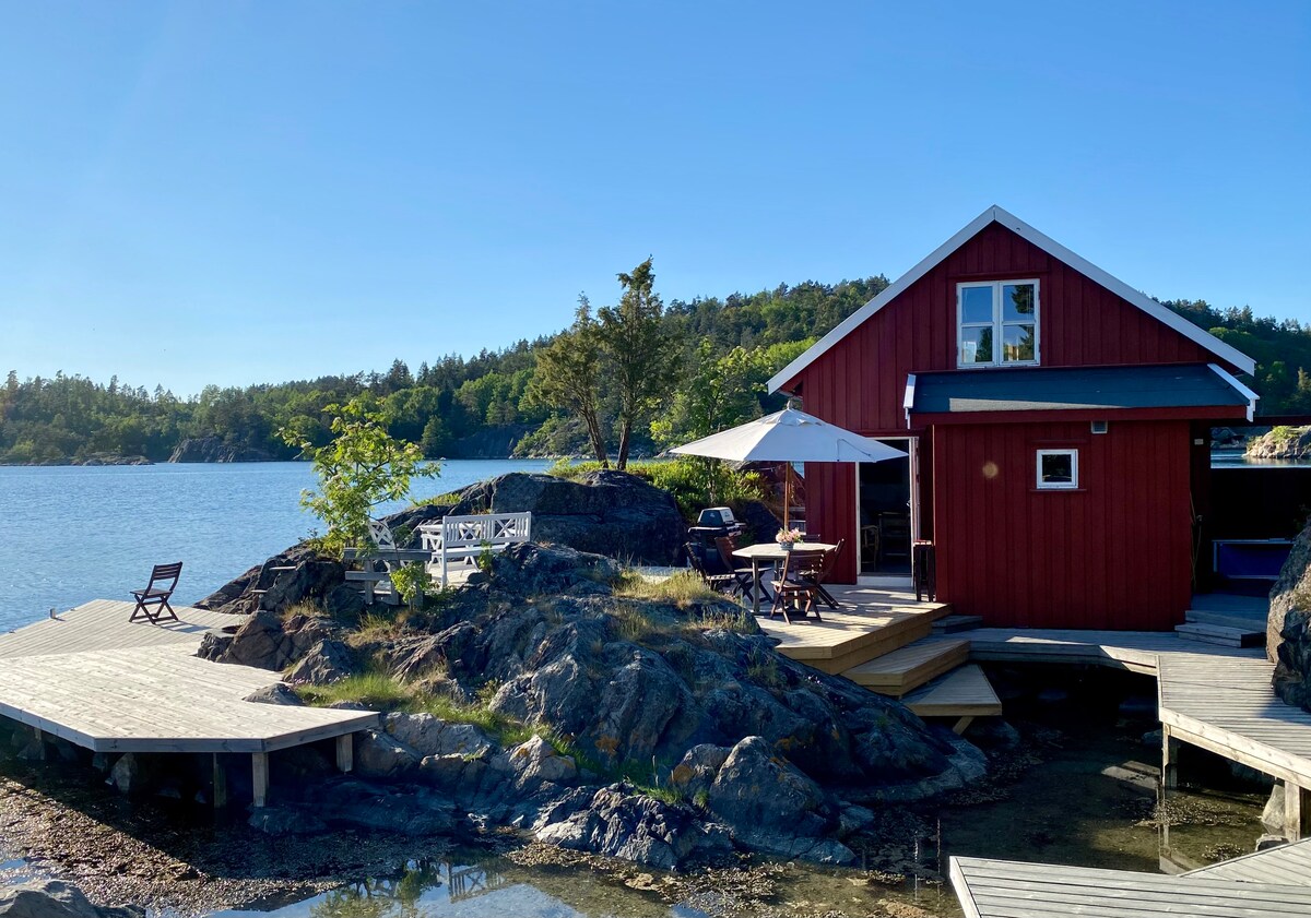 A charming red cottage is situated on the rocky shoreline, surrounded by greenery and water. Wooden decks extend towards the water, with outdoor seating areas highlighted by an umbrella. The serene landscape includes blue skies and distant hills, contributing to a peaceful environment.