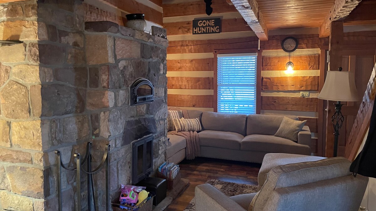 A cozy living area features a stone fireplace, inviting seating arrangement with a light grey sofa and a single armchair. Wooden accents and warm lighting create a welcoming atmosphere. A window with blinds allows natural light to fill the space.