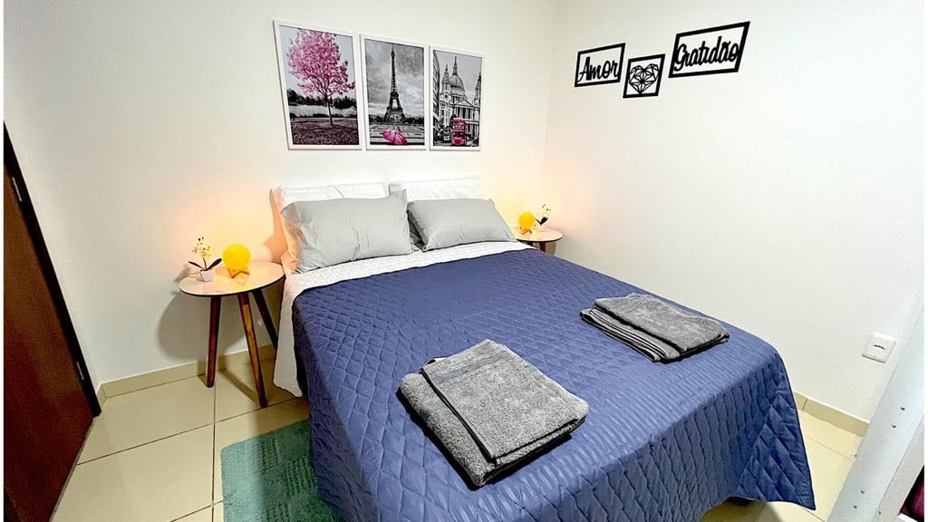 A spacious sleeping area features a large bed with gray pillows and a navy blue quilt. Two side tables with lamps are positioned beside the bed. Art pieces depicting urban scenes adorn the wall above, contributing to a serene ambiance.