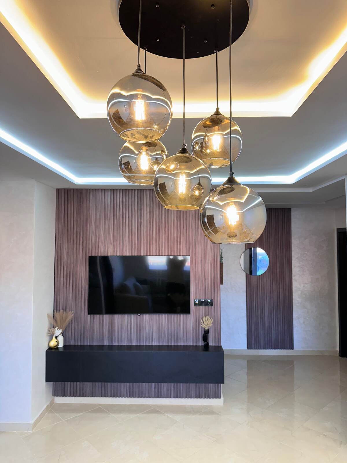 An elegantly designed living area features a sleek television mounted on a textured accent wall. Modern pendant lights hang from the ceiling, illuminating the space with a warm glow. A minimalist shelf below complements the contemporary decor.