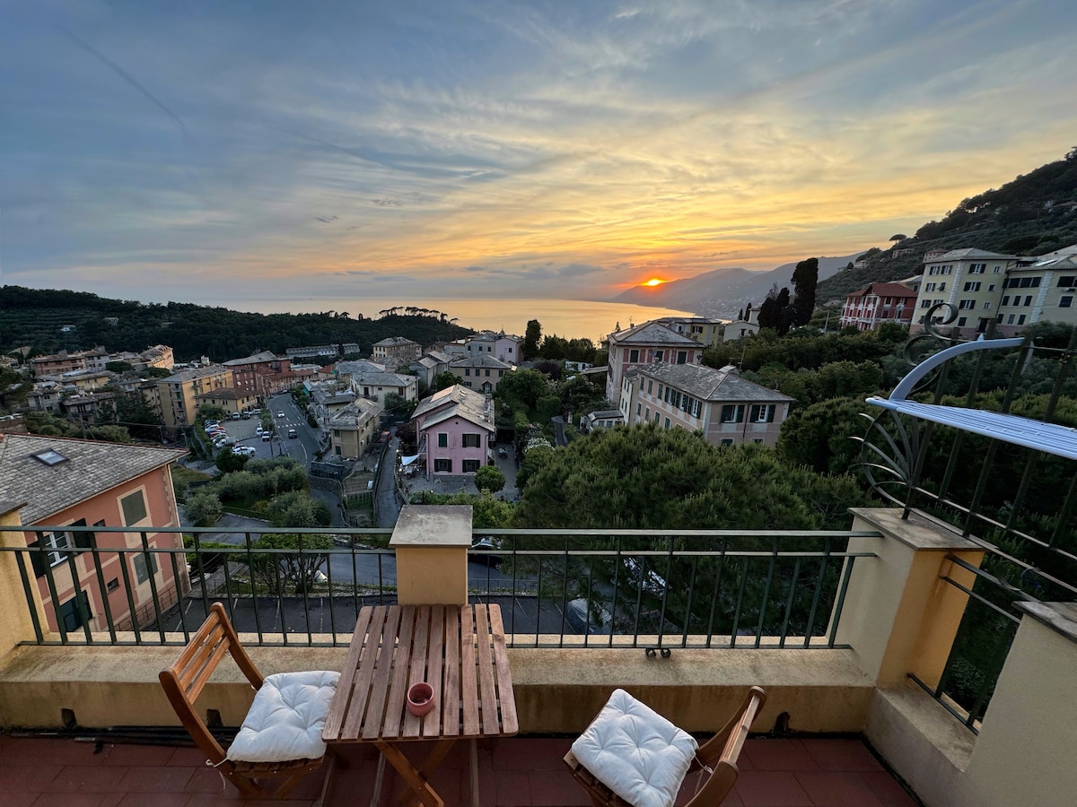 A terrace offers stunning sea views during sunset, framed by the surrounding landscape. A wooden table and two folding chairs create a cozy outdoor seating area, inviting moments of relaxation. The horizon is painted with warm hues, enhancing the scenic backdrop of nearby residences.